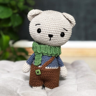 Amigurumi teddy bear wearing pants on a suspender and a green scarf.