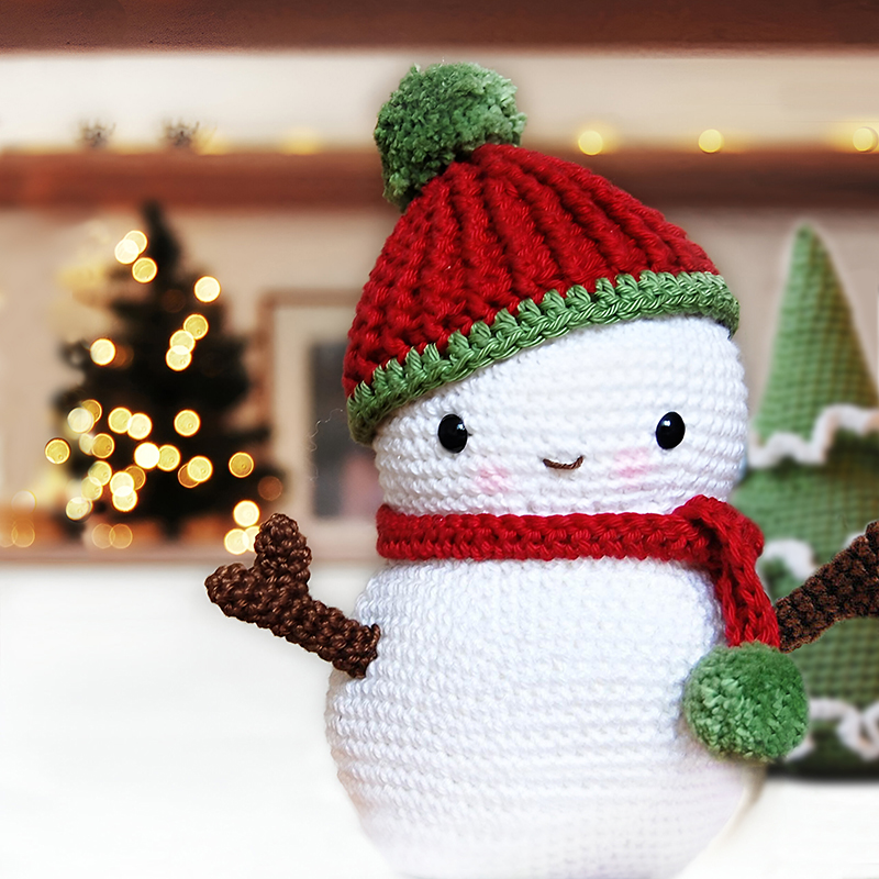 Amigurumi crochet snowman wearing a crocheted hat and scarf, adorned with pom poms. Behind the snowman is a crochet Christmas tree, and in the background, a real miniature Christmas tree is adorned with lights, softly blurred for a bokeh effect.