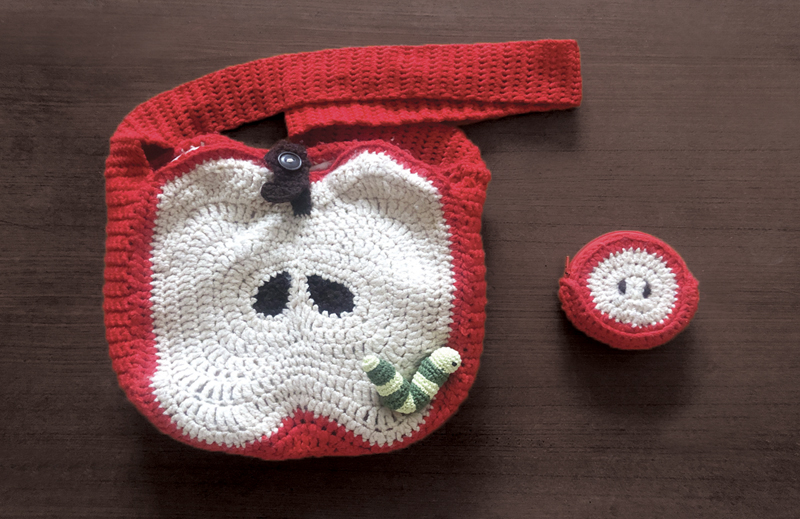 A crochet apple slice bag and a small, adorable apple wallet resting on a wooden desk. The bag features a realistic apple slice design with seeds and a small snail, while the wallet complements it with a charming mini apple motif. 