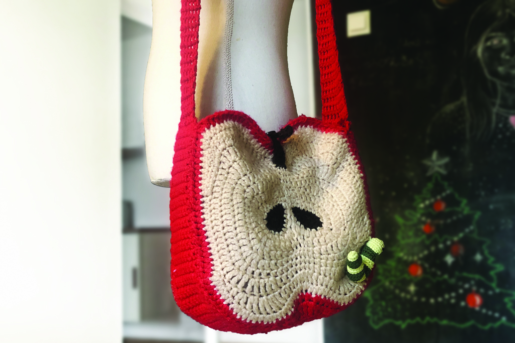 A handmade crochet apple slice bag with seeds and a leafy stem, hanging on a mannequin torso. A cute crochet snail is attached to the bag, adding a playful detail. The mannequin highlights the bag's size and shape, making it both stylish and functional—perfect for adding a handmade touch to any outfit!