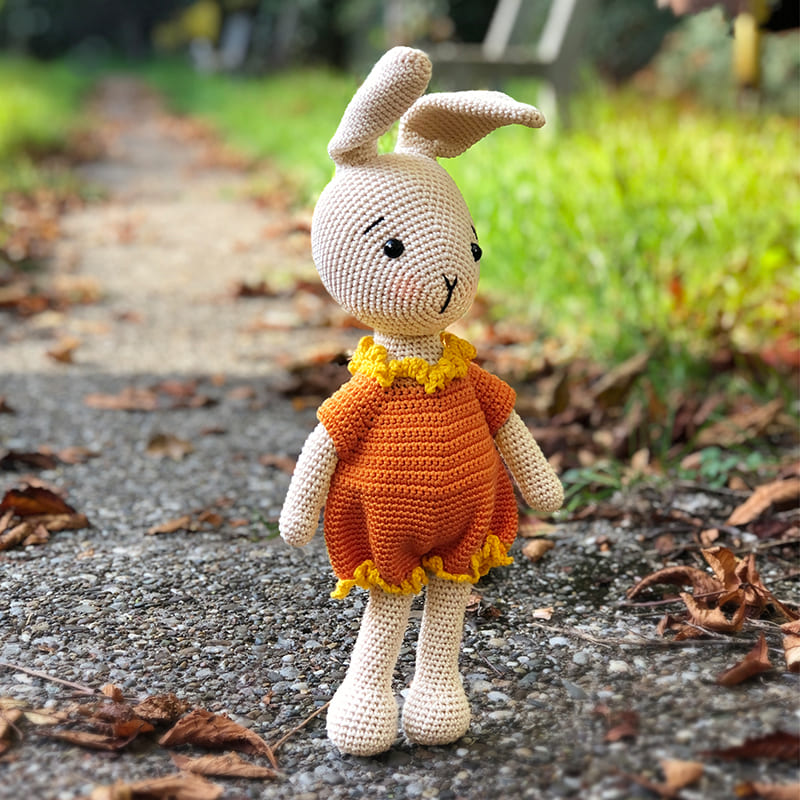 Cute amigurumi crochet bunny in a cozy yellow romper, standing among fallen leaves in a park.