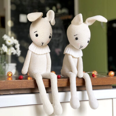 Two white crochet Easter bunnies sitting on a wooden table, with blurred flowers and a black chalkboard wall in the background. Perfect for Easter or spring decor.