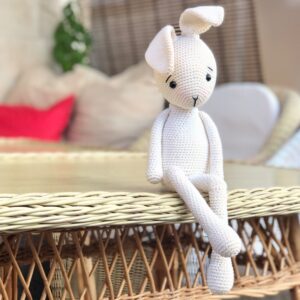 Crochet bunny sitting on the table in the backyard with crossed legs and red pillow behind.