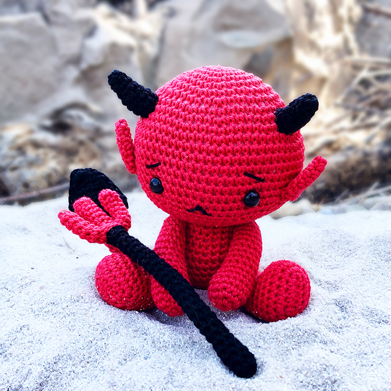 Cute crochet baby devil holding a pitchfork, sitting on the white sand and looking sad.