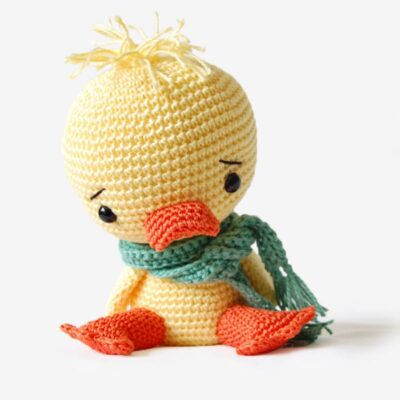 Crochet duck wearing green scarf sitting on the floor.