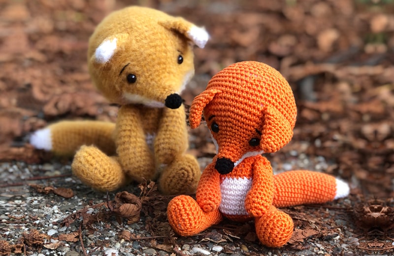 Two crochet foxes sitting in the forest with autumn leaves scattered around.