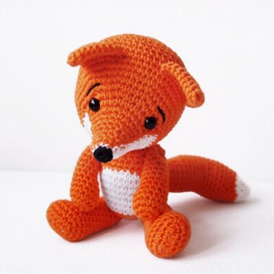 Crochet fox made in orange and white yarn, sitting on the floor.