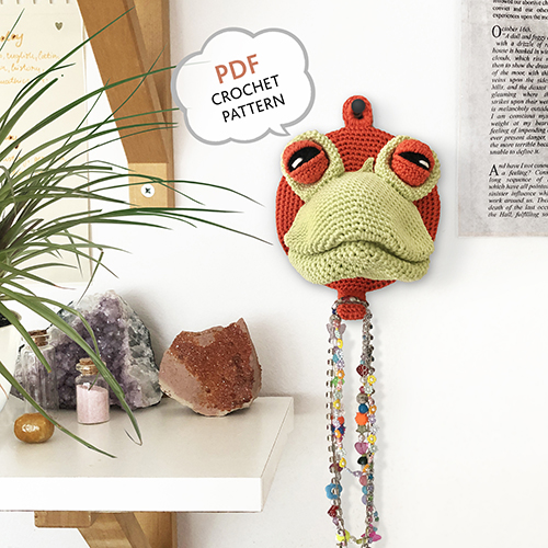 Crochet frog head mounted on the wall with necklaces hanging from it, adding a playful faux taxidermy charm to the decor.