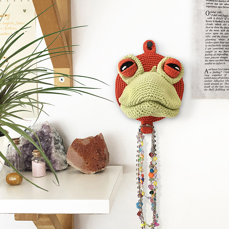 Crochet frog head mounted on the wall with necklaces hanging from it, adding a playful faux taxidermy charm to the decor.