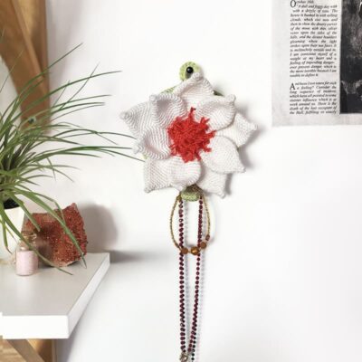 Crochet lotus flower wall mount hanging on the wall, with a necklace on its hook. To the left, there are crystals and a potted plant, while a poster is visible on the right side.