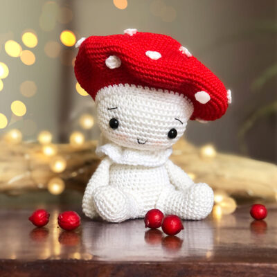 Crochet amigurumi mushroom doll resembling a red and white fly agaric (Amanita) sitting on a wooden table, surrounded by scattered red berries. A wooden branch and soft light in the background create a cozy atmosphere.