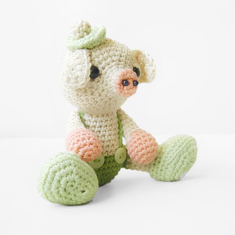 Crocheted pig sitting on the floor, wearing green suspenderpants and a light green berret.