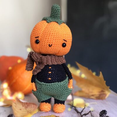 Crochet pumpkin doll dressed in green pants and a brown scarf, standing on a table. Autumn leaves and two pumpkins in the background enhance the Halloween atmosphere.