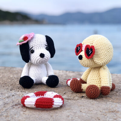 Two amigurumi dogs, one wearing a pink hat and the other with heart-shaped sunglasses, sitting on a concrete promenade by the sea with a lifebelt lying beside them.