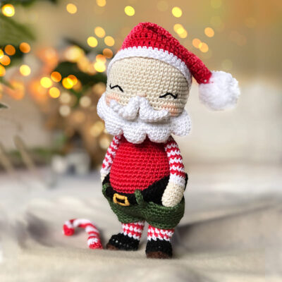 Handmade crochet Santa Claus doll standing with a candy cane beside it, with warm Christmas lights softly glowing in the background.