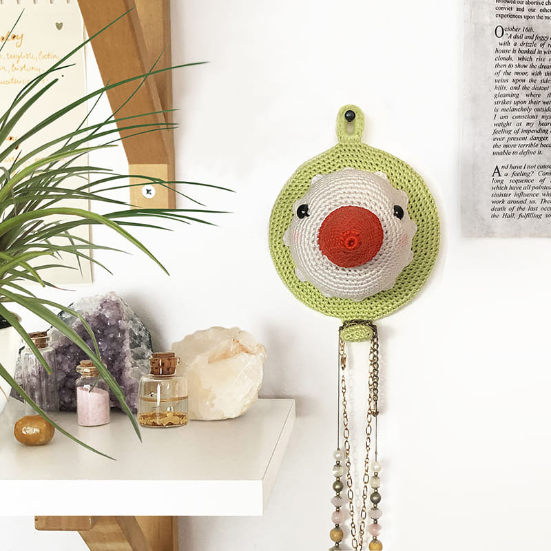 Crochet amigurumi stork head wall mount hanging on the wall, with a necklace on its hook. To the left, there are crystals and a potted plant.